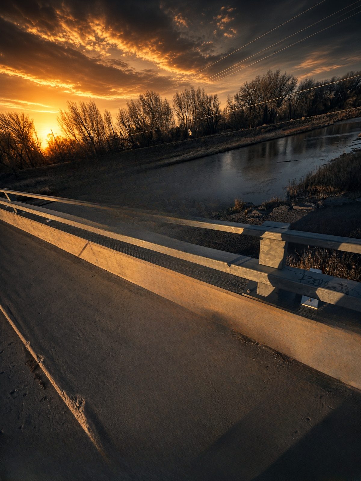 An empty bridge over a river at sunset, with bare trees and a glowing sky